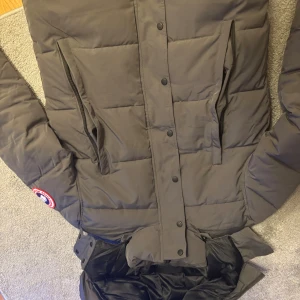 Mörkgrå dunjacka från Canada Goose - Säljer en mörkgrå dunjacka från Canada Goose med huva och stora fickor med dragkedja. Jackan har ett broderat märke på ärmen och knäppning med både dragkedja och knappar framtill. Insidan är fodrad i svart och jackan är perfekt för kalla dagar.