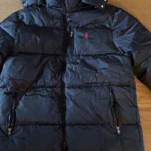Svart pufferjacka från Polo Ralph Lauren - Svart pufferjacka från Polo Ralph Lauren med röd broderad logga på bröstet. Jackan har huva, flera dragkedjeförsedda fickor och quiltad design. Materialet är syntet och dun, perfekt för kalla dagar. Stängs med dragkedja framtill och har snygga detaljer vid kragen.