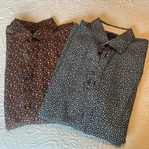 Två blommiga skjortor Selected Homme - Två slim fit skjortor från Selected Homme i storlek L. Ena skjortan är svart med vita små blommor, den andra har mörk botten med blommor i rött, gult och orange. Båda är i riktigt fint skick. 