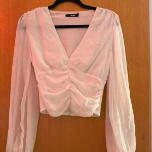 Vit omlottblus från Bik Bok - Säljer en vit omlottblus från Bik Bok i storlek S. Blusen har långärmade, transparenta ärmar och en snygg v-ringning. Framtill är den rynkad och croppad vilket ger en trendig look. Perfekt att styla med höga jeans eller kjol.