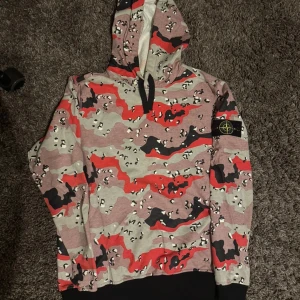 Camouflage hoodie från Stone Island - Säljer en fet hoodie från Stone Island med röd, svart och beige camouflage-mönster. Tröjan har huva med snörning, ribbade muddar och klassisk Stone Island patch på ärmen. Materialet är mjuk bomull, perfekt för chill dagar. Passar dig som vill sticka ut med streetwear.