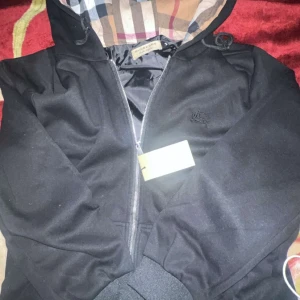 Svart vindjacka från Burberry - Snygg svart vindjacka från Burberry med klassiskt rutigt foder i huvan. Jackan har dragkedja framtill, ribbade muddar och en broderad Burberry-logga på bröstet. Perfekt för dig som vill ha en stilren och ikonisk jacka med street-vibe.