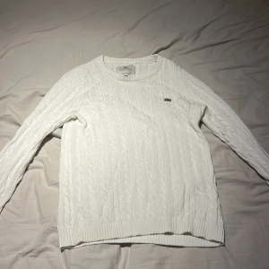 Vit kabelstickad tröja Lexington - Säljer en vit kabelstickad tröja från Lexington med rund hals och diskret logga på bröstet. Tröjan har långa ärmar och ribbade muddar vid ärmslut och nederkant. Perfekt för dig som gillar klassisk och clean stil.