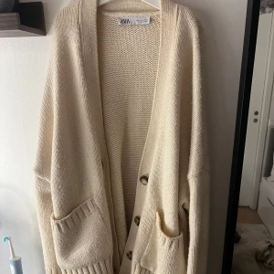 Beige stickad kofta från Zara - Oversized beige stickad kofta från Zara med långa ärmar, djupa fickor och knappar framtill. Koftan har bred ribbad kant vid ärmslut och nederkant, perfekt för lager på lager. Enkel och chill stil som funkar året runt.