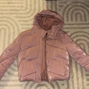Rosa pufferjacka från FB sister (New Yorker) - Säljer en snygg rosa pufferjacka från FB Sister i storlek S. Jackan har huva, stora fickor och dragkedja med tryckknappar. Materialet är glansigt och quiltat, perfekt för kalla dagar. Jackan har en croppad passform och cool look. (lite sönder vid fickan)