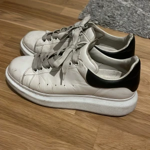 Vita sneakers från Alexander McQueen - Stilrena vita sneakers från Alexander McQueen med svart hälparti och tjock vit sula. Skorna har snörning och är tillverkade i skinn, vilket ger en lyxig känsla. Perfekta för dig som gillar en chunky och trendig look. Smutsen går enkelt att ta bort med lite vatten o gnug. Pris kan diskuteras såklart 