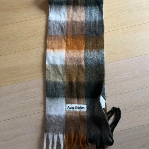 Rutig halsduk från Acne Studios - Snygg och fluffig halsduk från Acne Studios i rutigt mönster med färger som orange, brun, grå, vit och beige. Den är lång med fransar i ändarna och har en mjuk och lyxig känsla tack vare materialet i ull och mohair. Perfekt för att ge din outfit en cool touch.