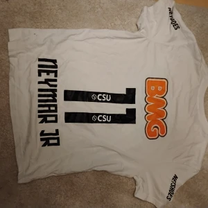 Vit Neymar Jr fotbollströja BMG - Vit fotbollströja med tryck på ryggen: Neymar Jr, nummer 11 och BMG-logga i orange. Svarta detaljer och sponsortryck på ärmarna. Rund hals och korta ärmar. Materialet är mjukt och lätt, perfekt för träning eller match.