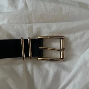 Svart bälte med guldfärgat spänne - Stilrent svart bälte med ett rektangulärt guldfärgat metallspänne och två matchande guldfärgade hällor. Bältet har en klassisk och enkel design som passar till jeans eller kostymbyxor. Materialet ser ut att vara skinn eller syntet.