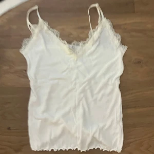 Vit linnetopp med spetsdetaljer - Säljer en söt vit linnetopp med smala axelband och v-ringad hals. Toppen har fina spetsdetaljer längs halsringningen och är tillverkad i mjukt ribbat material. Perfekt för dig som gillar en enkel men ändå romantisk stil.