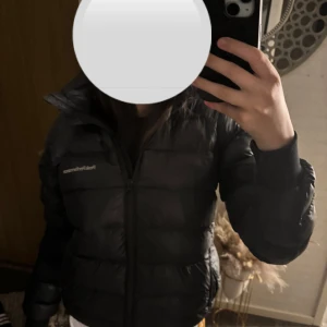 Svart Peak Performance dunjacka - Säljer en svart dunjacka från Peak Performance med huva och broderad logga på bröstet. Jackan har dragkedja framtill, ribbade muddar vid ärmslut och en puffad design som håller dig varm. Perfekt för kalla dagar och snygg stil.