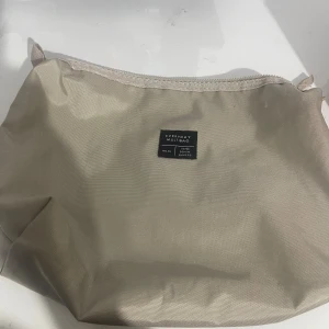 Beige Everyday Multibag med dragkedja - Snygg beige multibag från MENN MAKERS med stilren design och praktisk dragkedja upptill. Perfekt storlek för att organisera dina prylar och enkel att ta med sig. Väskan har en diskret etikett framtill med texten 'EVERYDAY MULTIBAG'.