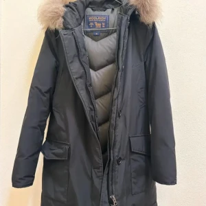 Woolrich - Parkajacka från Woolrich i svart med fluffig päls på huvan. Jackan har quiltad insida, långa ärmar med ribbade muddar, stora fickor och både dragkedja och knappar framtill. Perfekt för kalla dagar och riktigt snygg siluett.