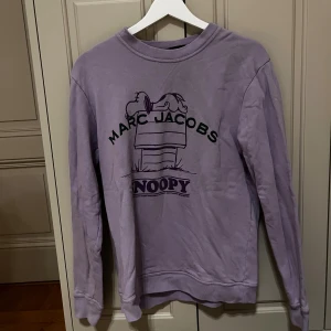 Lila Marc Jacobs x Peanuts sweatshirt XS - Superfin lila sweatshirt från Marc Jacobs x Peanuts med Snoopy-tryck på framsidan och ett stort statement-tryck på ryggen. Tröjan har rund hals, långa ärmar och är gjord i mjuk bomull. Perfekt för dig som gillar coola prints och comfy vibes.