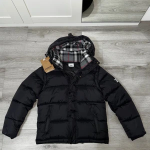 Svart pufferjacka + Vest Burberry   - Svart pufferjacka från Burberry med avtagbara ärmar, så du kan bära den som väst. Klassisk rutig huva i grått, vitt och rött. Jackan har dragkedja, tryckknappar och två fickor med zip. Burberry-logga på ärmen. Perfekt för kalla dagar. Ete 