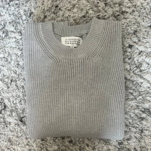 Maison Margiela tröja - Säljer en stilren grå stickad tröja från Maison Margiela. Tröjan har rund halsringning och ribbade muddar vid ärmslut och nederkant. Perfekt för lager-på-lager och enkel att matcha med olika outfits.