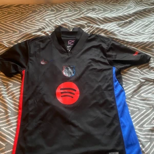 Svart FC Barcelona Nike fotbollströja L - FC Barcelona matchtröja från Nike i svart med röda och blå sidopaneler. Tröjan har korta ärmar, V-ringad krage och tryck med Spotify-logga samt klubbmärke på bröstet. Materialet är 100% polyester med Dri-Fit-teknologi för att hålla dig torr. UNHCR-tryck på ryggen.