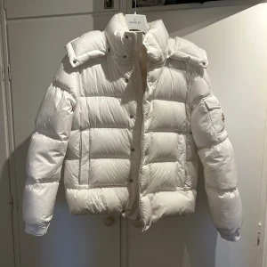 Vit Moncler jacka - Säljer en vit pufferjacka från Moncler med huva och hög krage. Jackan har Moncler-logga på ärmen, praktiska fickor och dragkedja framtill. Insidan har en cool serietidningsdetalj. Perfekt för dig som vill ha en clean och stilren vinterlook. Storlek 3 typ en medium lite större. Kvitto finns köpt på nk förra vinter