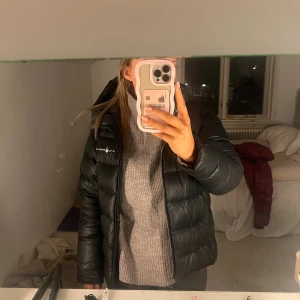 Svart pufferjacka från Save The Duck - Svart pufferjacka från Sailracing med huva och diskret logga på bröstet. Jackan har en klassisk quiltad design och dragkedja framtill. Perfekt för kalla dagar och har en snygg passform. Finns små defekter som inte påverkar jackans funktion (bilder finns). Tvättas innan den skickas.