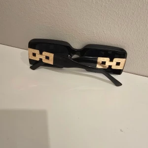 Svarta solglasögon med guldiga detaljer - Snygga svarta solglasögon med rektangulär form och breda skalmar. På skalmarna sitter stora, guldiga dekorativa länkar som ger en lyxig och trendig vibe. Perfekt accessoar för att lyfta din stil och sticka ut i mängden.