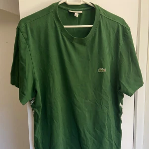 Grön Lacoste t-shirt med logga - Snygg grön t-shirt från Lacoste med klassisk krokodillogga på bröstet och vit/grön logoremsa längs sidan. Rund hals och korta ärmar. Perfekt för dig som gillar sportig och stilren design. Materialet är mjuk bomull som känns skönt mot huden.