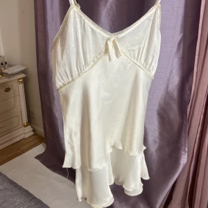 Vit,Crème satin Linne fr. Tessie.S-M - Supersöt vit kortklänning från Tessie i glansig satin med tunna axelband och volangkant. Klänningen har en liten rosett framtill och spetsdetaljer längs urringningen. Perfekt för dig som gillar en feminin och chic stil.Vit Crème färgad Linne