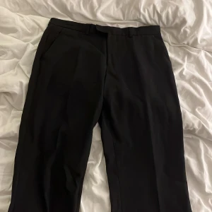 Svarta kostymbyxor från Steel, linne - Svarta kostymbyxor från Steel med klassisk rak passform. Byxorna har bälteshällor, sidfickor och två bakfickor med knapp. Materialet är 55% linne och 45% polyester, vilket ger en snygg och lite lyxig känsla. Perfekt för dig som gillar stilrena och enkla plagg.