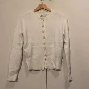 Vit kofta med pärlknappar från Zara - Superfin vit kofta från Zara med långa ärmar och rund hals. Koftan har unika pärlknappar som ger en lyxig touch och är stickad i ett mjukt material. Perfekt att slänga över en topp för en clean och stilren look.