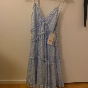 Blommig blå långklänning från NA-KD - Supersöt klänning från NA-KD med blommigt mönster och volangdetaljer. Klänningen har smala axelband och omlott-v-ringning, 