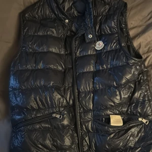 Mörkblå dunväst från Moncler - Snygg mörkblå dunväst från Moncler med glansig finish och klassisk logga på bröstet. Västen har hög krage, två dragkedjefickor framtill och quiltad design. Perfekt för lager-på-lager och ger en clean streetstyle-vibe.