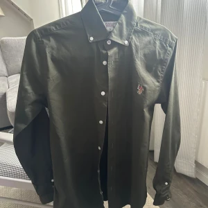 Mörkgrön skjorta från U.S. Polo Assn. - Snygg mörkgrön skjorta från U.S. Polo Assn. i storlek S. Skjortan har klassisk krage med vita knappar, lång ärm och broderad logga på bröstet. Tillverkad i mjuk bomull och har en liten detalj med röd, vit och blå rand vid manschetten.