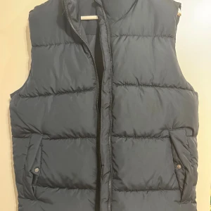 Svart dunväst från C, storlek S - Svart dunväst från C i storlek S. Västen har en quiltad design med två fickor framtill som stängs med tryckknappar och en dragkedja i mitten. Perfekt att slänga över en hoodie för extra värme under höst och vinter. Har haft på mig den en gång 