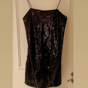 Svart paljettklänning med smala axelband från H&M  - Supersnygg svart kort klänning med glittriga paljetter över hela ytan och smala axelband. Klänningen har en rak och enkel siluett som ger en cool vibe. Perfekt för dig som vill sticka ut med lite extra bling. Fodrad insida för skönare känsla.