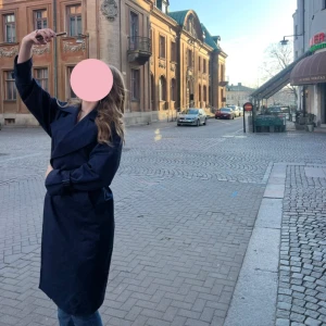 Mörkblå trenchcoat med bred krage - Snygg mörkblå trenchcoat med bred krage och långa ärmar. Jackan har en klassisk omlott-design och är tillverkad i ett slätt material som känns lätt och bekvämt. Perfekt för dig som gillar stilrena och tidlösa plagg med lite edge.