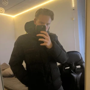 Svart pufferjacka från Superdry S - Svart pufferjacka från Superdry med broderad logga på bröstet och huva. Jackan har en clean look med diskreta detaljer i orange och stängs med dragkedja och knappar. Perfekt för kalla dagar och riktigt skön passform.