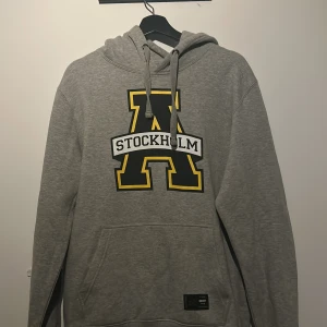 Grå hoodie med AIK Stockholm-tryck - Säljer en grå hoodie med stort A och Stockholm-tryck i svart och gult. Tröjan har en klassisk design med dragsko i huvan och en praktisk magficka. Perfekt för en avslappnad stil.