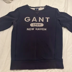 Mörkblå sweatshirt från GANT - Snygg mörkblå sweatshirt från GANT med tryck 'GANT 1949 NEW HAVEN' på bröstet. Klassisk rund halsringning och långa ärmar. Perfekt för dig som gillar stilren och sportig look. Mjuk och skön bomullsblandning.