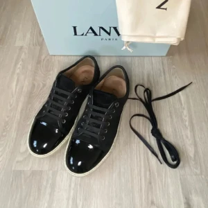 Svarta sneakers från Lanvin  - Snygga svarta sneakers från Lanvin med blank tå i lack och sidor i mocka. Skorna har rund tå, snörning och vit platt sula. Perfekt för dig som gillar stilrena och exklusiva sneakers med en cool kontrast mellan material. (Endast skorna ingår)