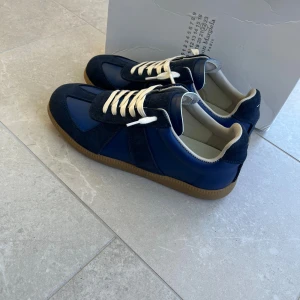 Blå Maison Margiela sneakers - Maison Margiela sneakers i mörkblått med vita snören och beige insida. Ovandel i skinn och mocka, gummisula och rund tå. Klassisk sportig siluett med snygga kontraster mellan material och färger. Perfekt för dig som gillar stilrena och unika sneakers.
