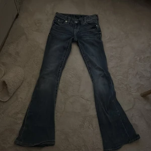 True Religion bootcut jeans blå, stl 24 - Säljer ett par blå bootcut jeans från True Religion i storlek 24. Jeansen har tydliga kontrastsömmar, klassiska fickor med lock och knappar bak samt broderade detaljer. Tillverkade i jeansmaterial med snygg tvättad look.