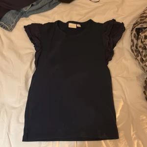 Mörkblå topp med volangärmar Gina Tricot - Snygg mörkblå topp från Gina Tricot med korta volangärmar som ger en cool och feminin touch. Rund halsringning och rak passform. Perfekt för dig som gillar stilrena plagg med en twist. Materialet känns mjukt och lätt mot huden.