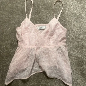 Ljusrosa broderad linnetopp Hollister - Supersöt ljusrosa linnetopp från Hollister med tunna axelband och v-ringad hals. Toppen har broderade detaljer och är tillverkad i ett lätt bomullstyg. Perfekt för varma dagar och ger en romantisk vibe till din outfit.