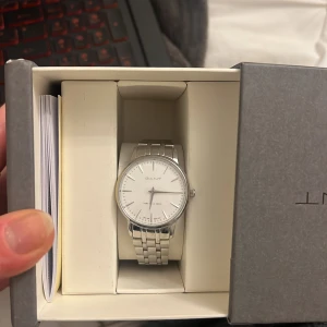 Silverfärgad klocka från GANT - Äkta stilren klocka från GANT med rund vit urtavla och silverfärgat armband i metall. Klockan har minimalistisk design med tydliga markeringar och GANT-logga på både urtavla och spänne. Levereras i originalask. 