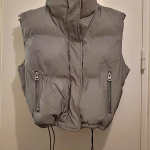 Grå dunväst, reflex, från FB Sister XS - Säljer en grå dunväst från FB Sister i storlek XS. Västen har hög krage, två fickor med dragkedja och snörning nedtill för att justera passformen. Perfekt för lager-på-lager och har en clean, modern look. Liten defekt som finns på bild.