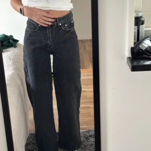 Svarta  raka jeans - Säljer ett par svarta jeans med hög midja och raka, lite bredare ben. Klassisk femficksmodell i kraftig denim med silverfärgade knappar och detaljer. Perfekta för dig som gillar en avslappnad och tidlös look.