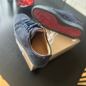 Christian Louboutin Skor Mörkblå  - Snygga mörkblå sneakers från Christian Louboutin med klassisk röd sula och rund tå. Skorna är tillverkade i mocka med skinnfoder och har snörning framtill. Perfekt för dig som vill ha en lyxig och stilren look med exklusiva detaljer.