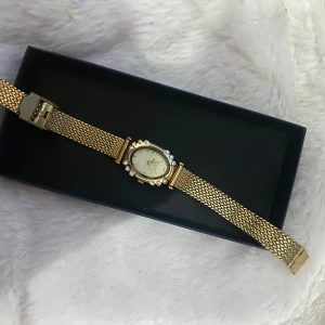 Guldfärgad klocka med pärlor - Säljer en elegant guldfärgad klocka med oval urtavla och mesh-armband. Runt urtavlan sitter glittrande pärlor som ger extra lyxig känsla. Perfekt accessoar för dig som gillar stilrena detaljer och guldtoner.