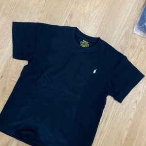 Svart t-shirt från Polo Ralph Lauren - Svart t-shirt från Polo Ralph Lauren med klassisk vit broderad logga på bröstet. Modellen är slim fit och har rund hals samt korta ärmar. Tillverkad i mjuk bomull som känns skön mot huden. Perfekt för dig som gillar stilrena och tidlösa plagg.