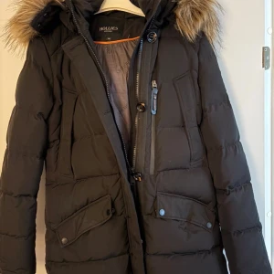 Svart lång dunjacka Hollies med päls ÄKTA - Säljer en svart ÄKTA dunjacka från Hollies med fluffig fuskpäls på huvan. Jackan har långa ärmar, flera fickor med knappar och dragkedja framtill. Den är quiltad och har en snygg, klassisk look som passar perfekt när det är kallt ute.