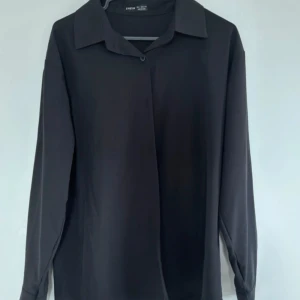 Svart långärmad skjorta från Shein - Stilren svart skjorta från Shein med klassisk krage och lång ärm. Skjortan har en enkel design utan synliga knappar framtill och är tillverkad i ett mjukt, lätt material som ger en snygg och clean look.